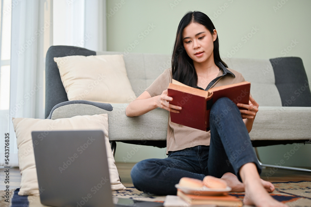 Attractive Asian woman reading a book, doing her college homework in living room. Attractive Asian woman reading a book, doing her college homework in living room.