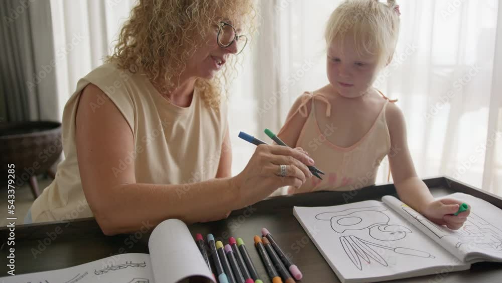 Grandmother plays with her granddaughter and teaches her to draw with bright pencils in a coloring book, sitting in the living room. The concept of home education and togetherness with the family. Grandmother plays with her granddaughter and teaches her to draw with bright pencils in a coloring book, sitting in the living room. The concept of home education and togetherness with the family.