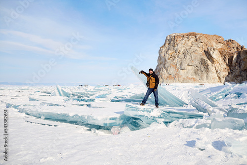 A man stands by a huge block of clear blue ice. Frozen Lake Baikal on a winter day. Ice blocks, hummocks covered with snow. Winter travel, active recreation. photo