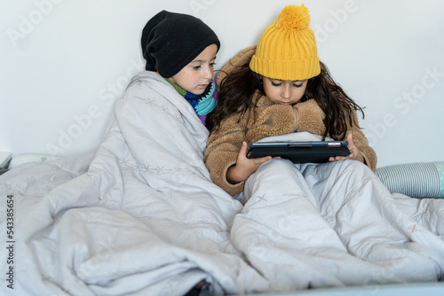 Children in warm clothes at home in cold winter time. Kids in faux fur coat, knitted hat and scarf sitting in a bed in home, warms in blanket and using tablet.