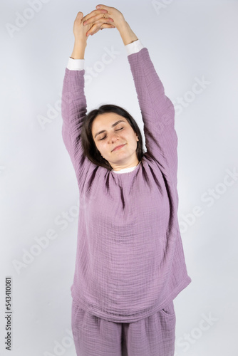 Young brunette woman in muslin lavender pajamas sleep clothes stretches by raising her arms up and closing her eyes