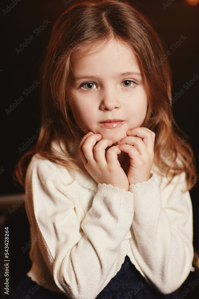 Little adorable smiling girl is touching face with her hands and looking in camera with a dark background. Child in lace blue dress and knit sweater is sitting on metal chair. Happy childhood Little adorable smiling girl is touching face with her hands and looking in camera with a dark background. Child in lace blue dress and knit sweater is sitting on metal chair. Happy childhood