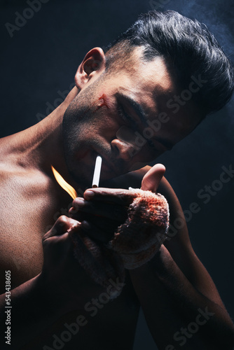 Boxer, man and cigarette smoking after fight, blood and bandage from injury against a black studio background. Fighter smoke after boxing, training or mma match with bruise, violence and blood scar