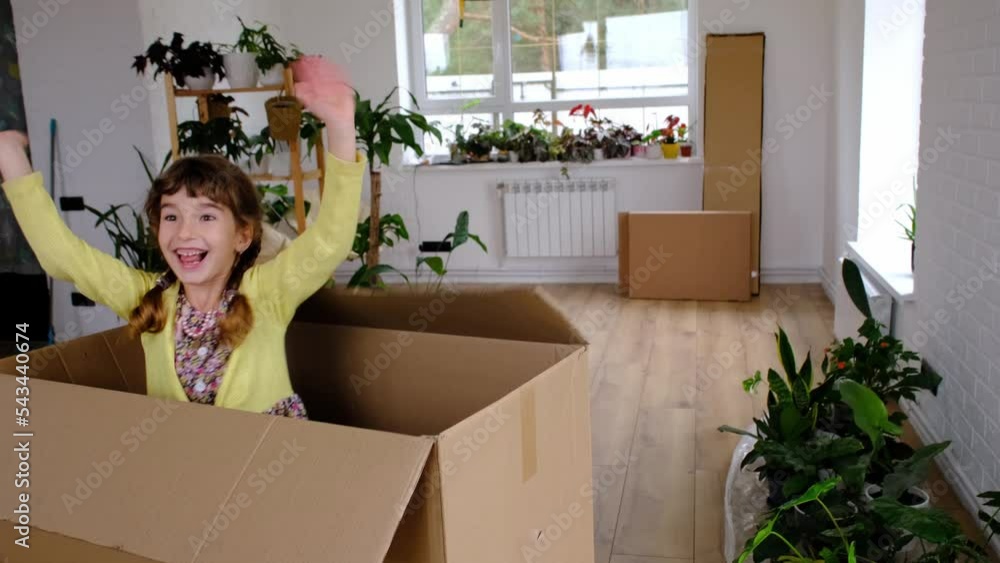 The girl hides in a big box and looks out of it funny in a room inside the home. Moving to a new house, unpacking things, renting an apartment, mortgage, housing issue The girl hides in a big box and looks out of it funny in a room inside the home. Moving to a new house, unpacking things, renting an apartment, mortgage, housing issue