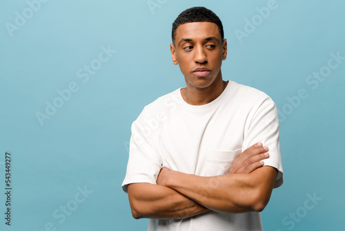 Pensive handsome serious young african-american guy wearing white casual t-shirt posing with folded hands isolated on blue background