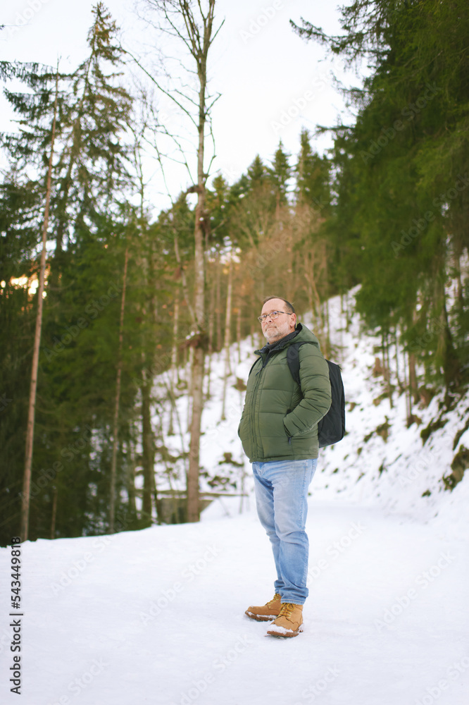 Outdoor portrait of middle age 55 - 60 year old man hiking in winter forest, wearing warm jacket and black backpack