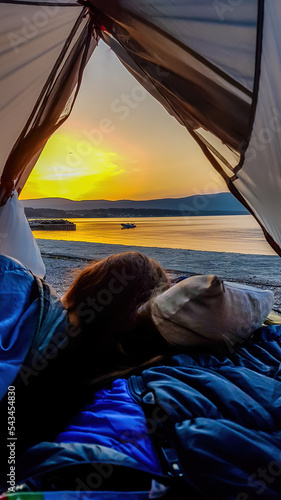 A girl lying in a tent set on the beach and watching the beautiful sunrise in Krk, Croatia. Sun is rising from behind the hill, turning sky orange. Calm surface of the sea reflects the sunbeams. © Chris
