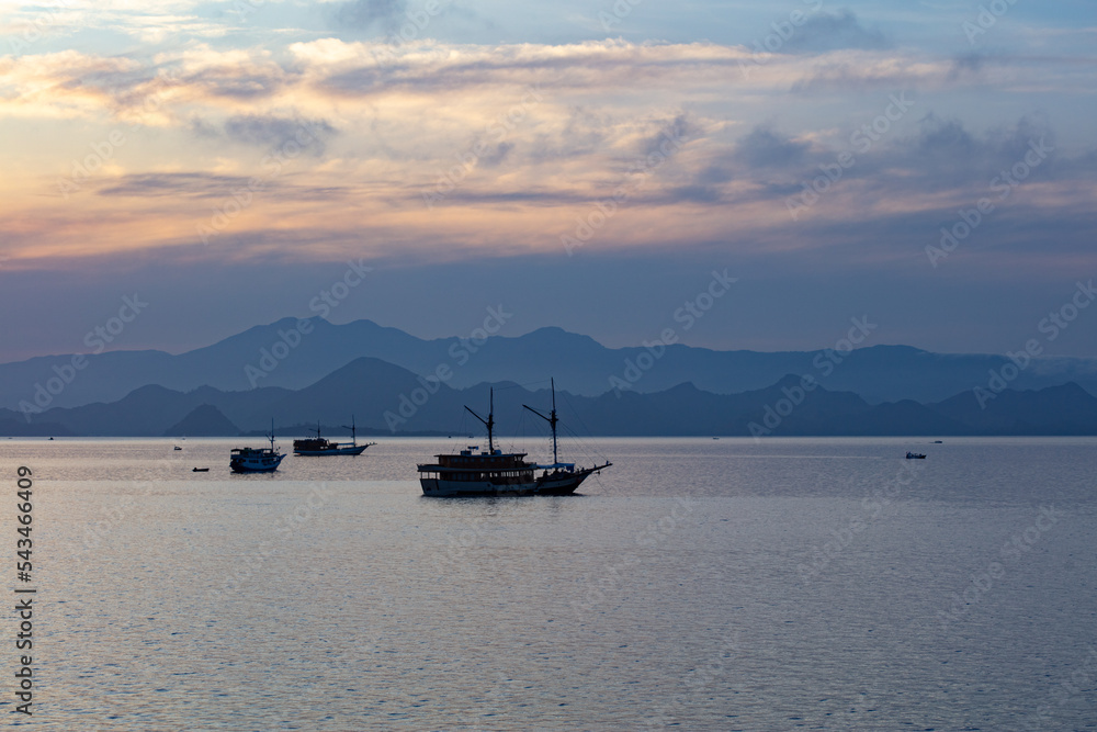 A tranquil sunrise silhouettes small boats in Komodo National Park, Indonesia. This beautiful park is popular for scuba diving, snorkeling, and for viewing Komodo dragons. A tranquil sunrise silhouettes small boats in Komodo National Park, Indonesia. This beautiful park is popular for scuba diving, snorkeling, and for viewing Komodo dragons.
