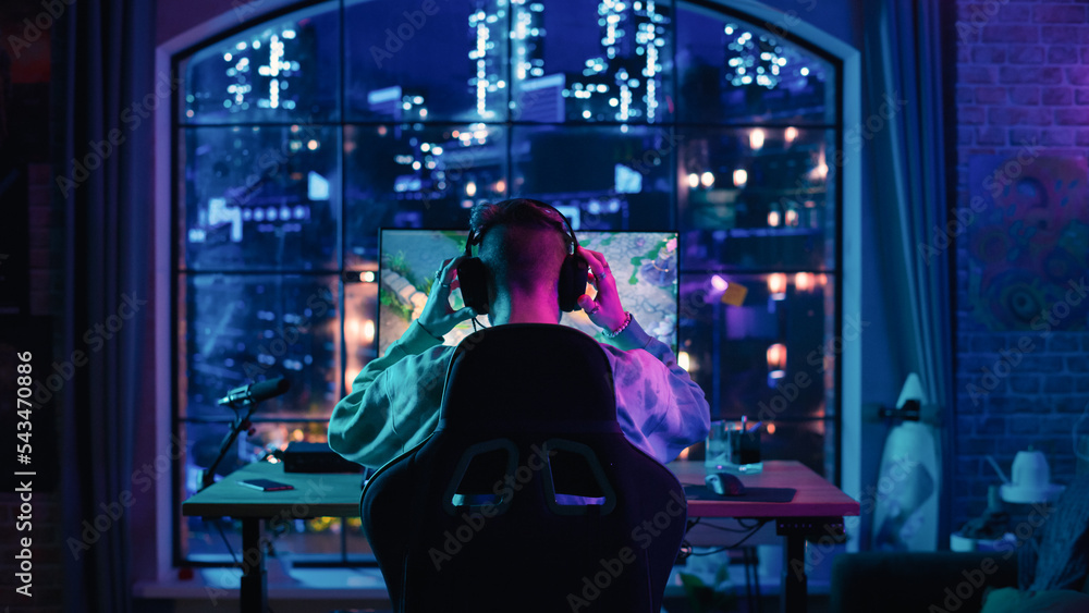 Back of the Head Angle of a Successful Stylish Gamer Putting on Headphones and Playing a Video Game on Personal Computer in a Neon Lit Living Room at Home. Young Man in Victorious.
