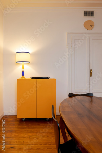 Detail yellow sideboard in kitchen with lighted lamp and wooden table with black chairs. Antique apartment interior with modern furniture