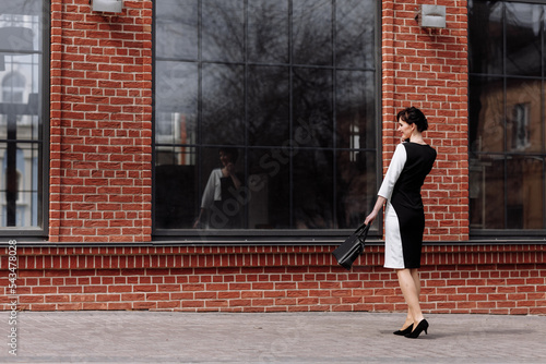 Happy lawyer businesswoman professional is walking outdoors talking on cell smart phone. Caucasian successful female is smiling wearing stylish white-black dress with black bag