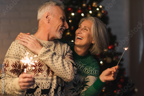 Canvas Print positive middle aged woman hugging bearded husband while holding bright sparkler