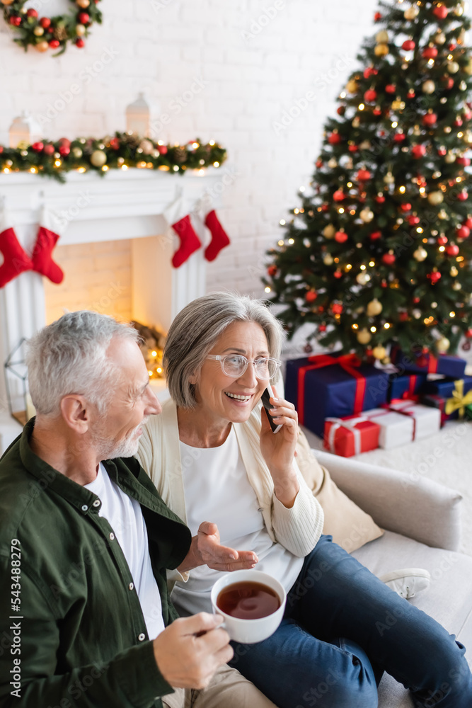 middle aged man holding cup of tea and looking at wife talking on smartphone near christmas tree middle aged man holding cup of tea and looking at wife talking on smartphone near christmas tree
