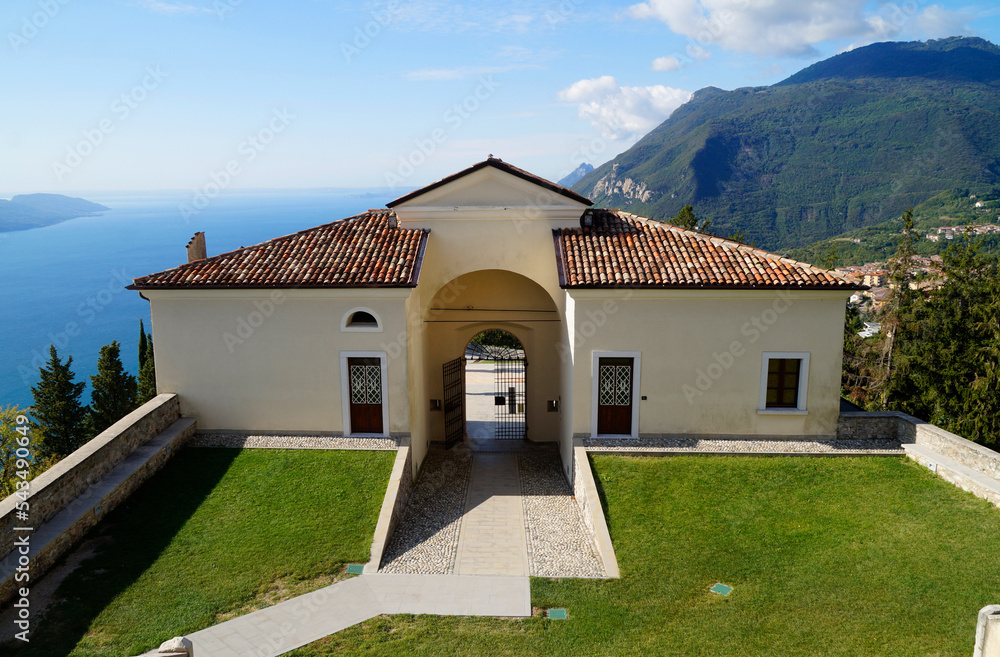 Fototapeta premium Mediterranean church Madonna di Montecastello in Tignale overlooking vast blue lake Gardasee with the Italian Alps against the clear blue sky in the background, Northern Italy, Lombardy