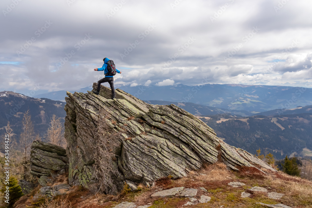 Man with a hiking backpack on a massive rock on summit Gertrusk on the way near Ladinger Spitz, Saualpe, Lavanttal Alps, Carinthia, Austria Europe. Trekking on cloudy early spring day. Wanderlust