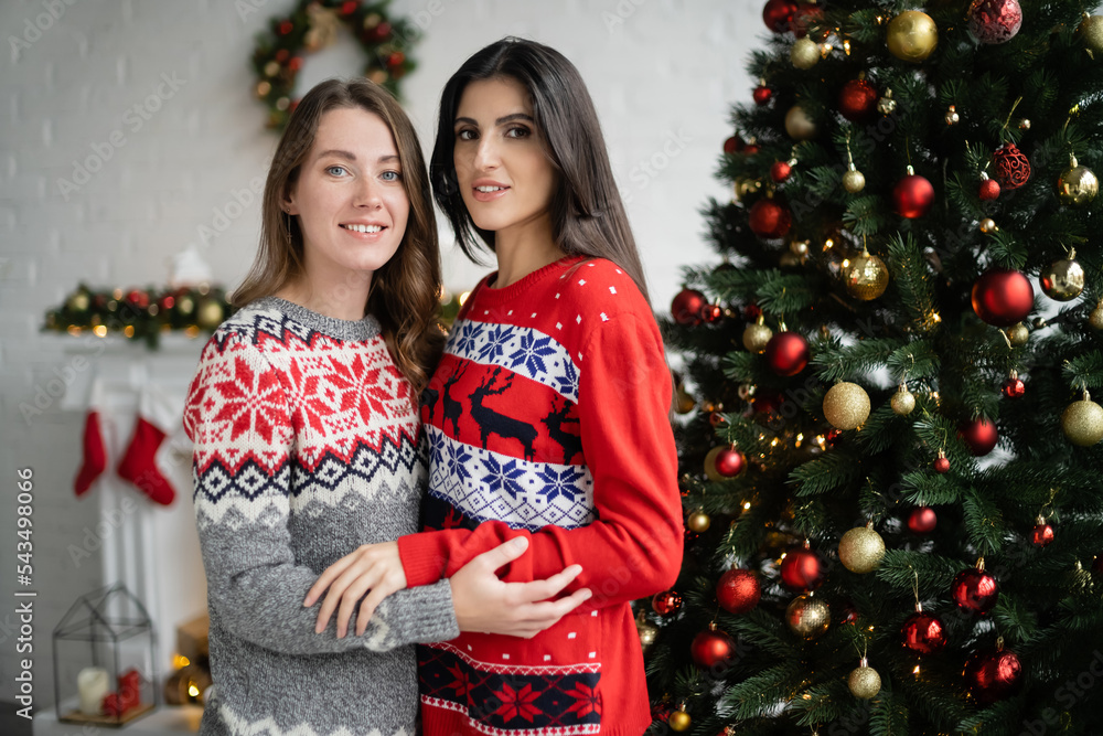 Smiling same sex couple in sweaters hugging and looking at camera near christmas tree Smiling same sex couple in sweaters hugging and looking at camera near christmas tree