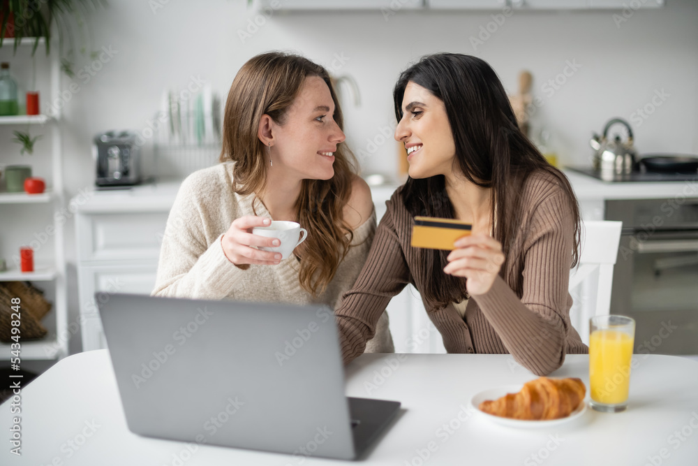 Side view of smiling same sex couple holding credit card and coffee near laptop and breakfast in kitchen Side view of smiling same sex couple holding credit card and coffee near laptop and breakfast in kitchen