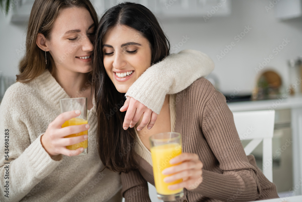 Young woman in sweater hugging girlfriend and holding orange juice in kitchen Young woman in sweater hugging girlfriend and holding orange juice in kitchen