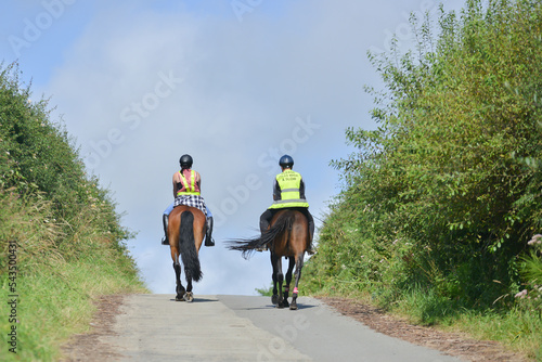 Riding over the hill, two horse riders riding their horses over the brow of a hill off on adventures with their horses on a summers day, wearing safety gear that makes them visible to other road users photo