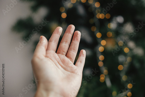 Merry Christmas and Happy Holidays! Hand in christmas golden glitter on festive background with golden lights. Atmospheric winter time. Person decorating room