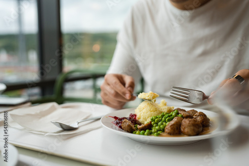Close up on hands of unknown caucasian man sit at the table with fork and knife eat meatballs with peas and mashed potato