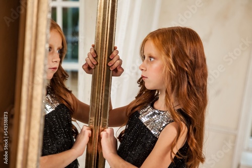 Little girl 5-6 years old with red hair, looking reflection, posing, fashion model in stylish elegant black dress in living room at mirror. Fashionable young lady actress at home. Copy text space