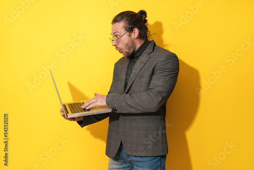 Surprised man wearing eyeglasses is using laptop computer on yellow background