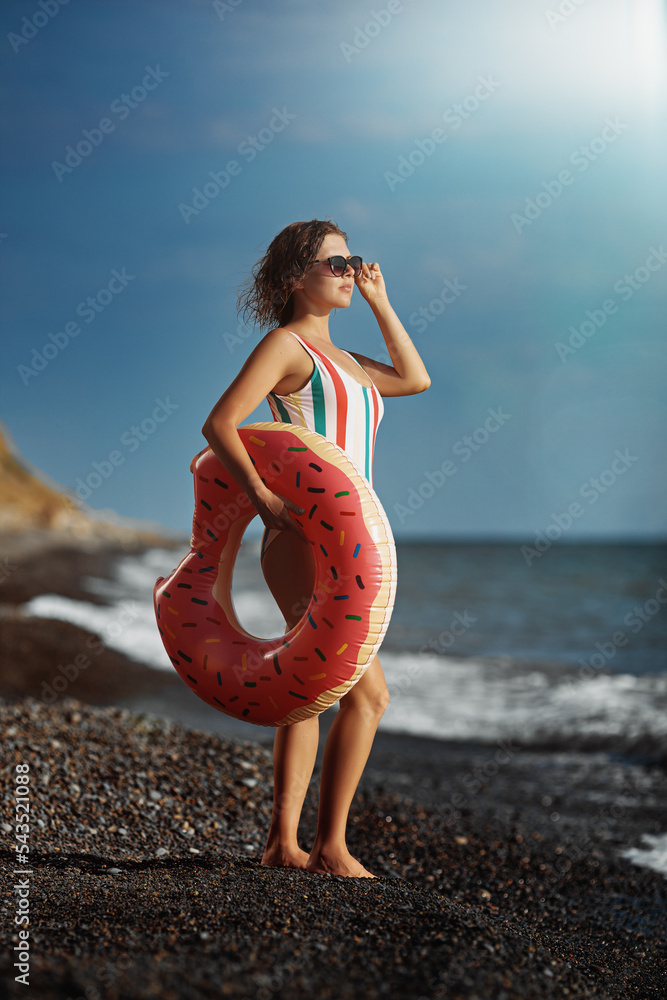 Tanned young woman in a swimsuit and sunglasses standing on a beach with an inflatable donut.