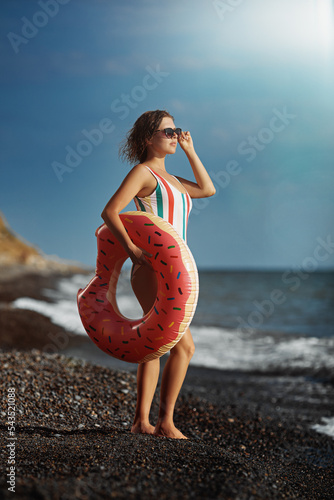 Obraz na płótnie Tanned young woman in a swimsuit and sunglasses standing on a beach with an inflatable donut. w salonie