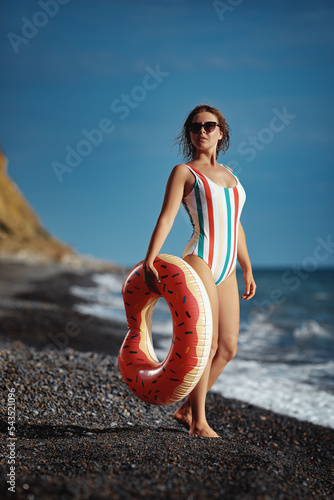 Obraz na płótnie Beautiful young woman in a swimsuit and sunglasses standing on a beach with an inflatable donut. w salonie