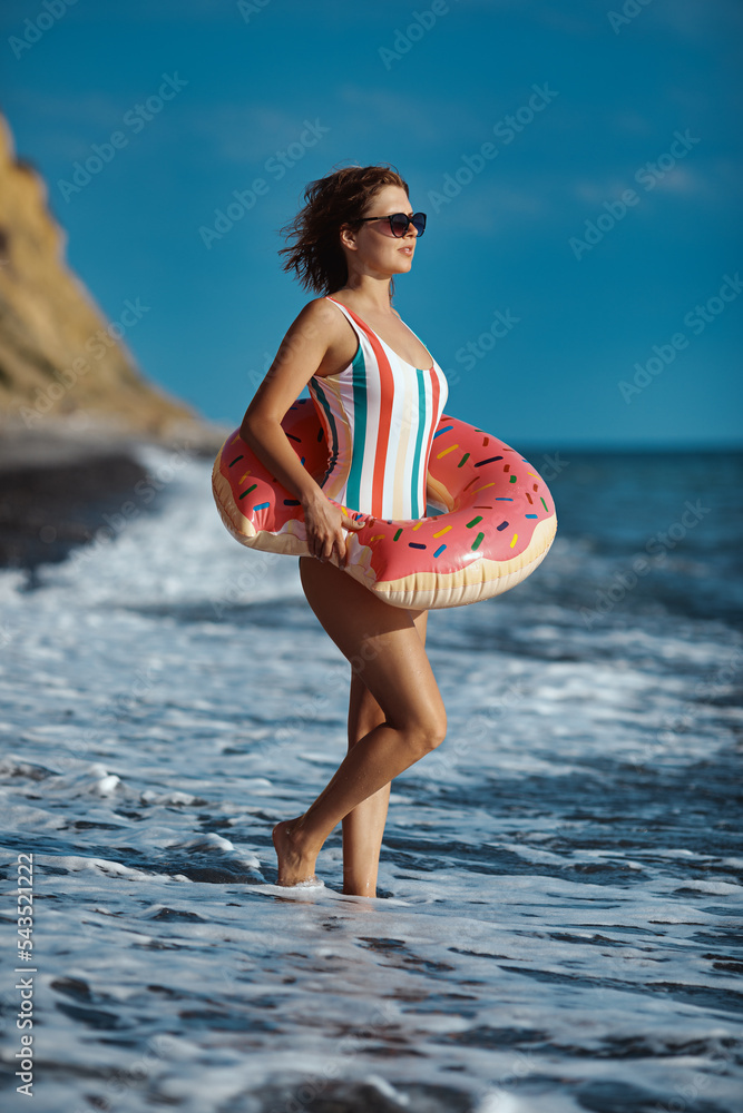 Tanned young woman in a swimsuit and sunglasses standing on a beach with an inflatable donut ring and looking at the see.