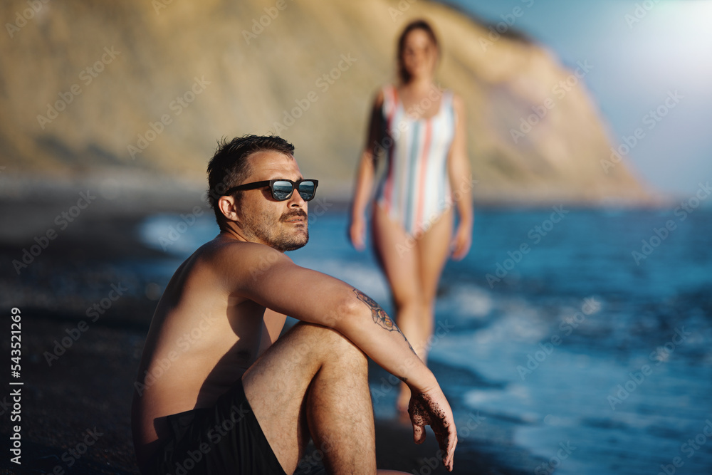 Young couple on the beach. Focus on the man looking at the see in the foreground. Pretty woman in the background. Shallow depth of field.