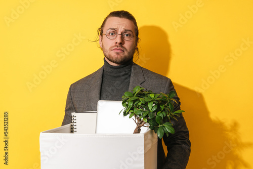 Upset man holding box with personal items after job resignation against yellow background