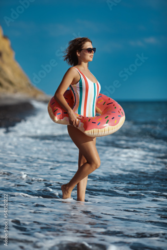Obraz na płótnie Tanned young woman in a swimsuit and sunglasses standing on a beach with an inflatable donut ring and looking at the see. w salonie