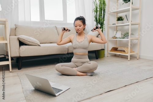Asian woman at home working out stretching and fitness while holding dumbbells online while looking at her laptop, home workouts to maintain a healthy body as a lifestyle
