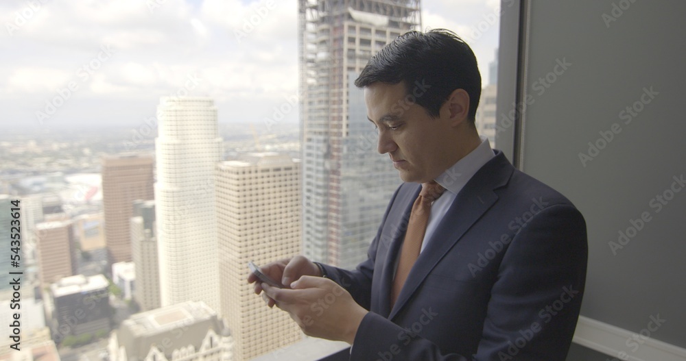 Successful young business man using internet on a smart phone in front of a skyscraper window with views across Los Angeles. Medium close up, side view Successful young business man using internet on a smart phone in front of a skyscraper window with views across Los Angeles. Medium close up, side view
