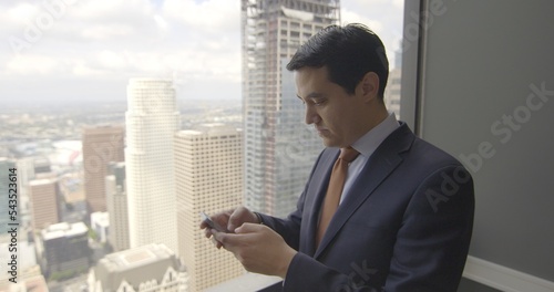 Successful young business man using internet on a smart phone in front of a skyscraper window with views across Los Angeles. Medium close up, side view