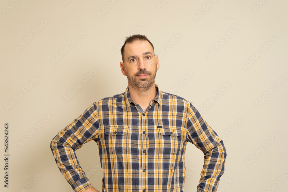 Fototapeta premium Cheerful bearded man in checkered shirt posing isolated on beige background, studio portrait. People lifestyle concept. Mock up copy space. Standing with your arms akimbo on your waist blinking.
