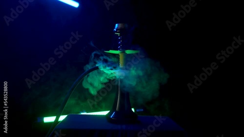 Stylish hookah with yellow shaft stands on dark table. White thick clouds of smoke rise from hot coals dissipating around hookah. Smoking hookah in bar