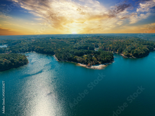 Fototapeta Naklejka Na Ścianę i Meble -  Lake Lanier in North Georgia, 4K aerial drone on a sunny fall day. Radiant clouds both blue and orange. Clear view of fall colored trees and blue lake water.