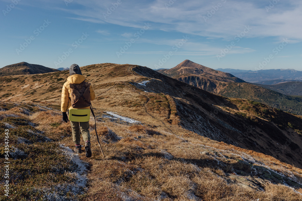 Woman hiker in yellow jacket with backpack climbs autumn mountains with light snow on ground on sunny cold day