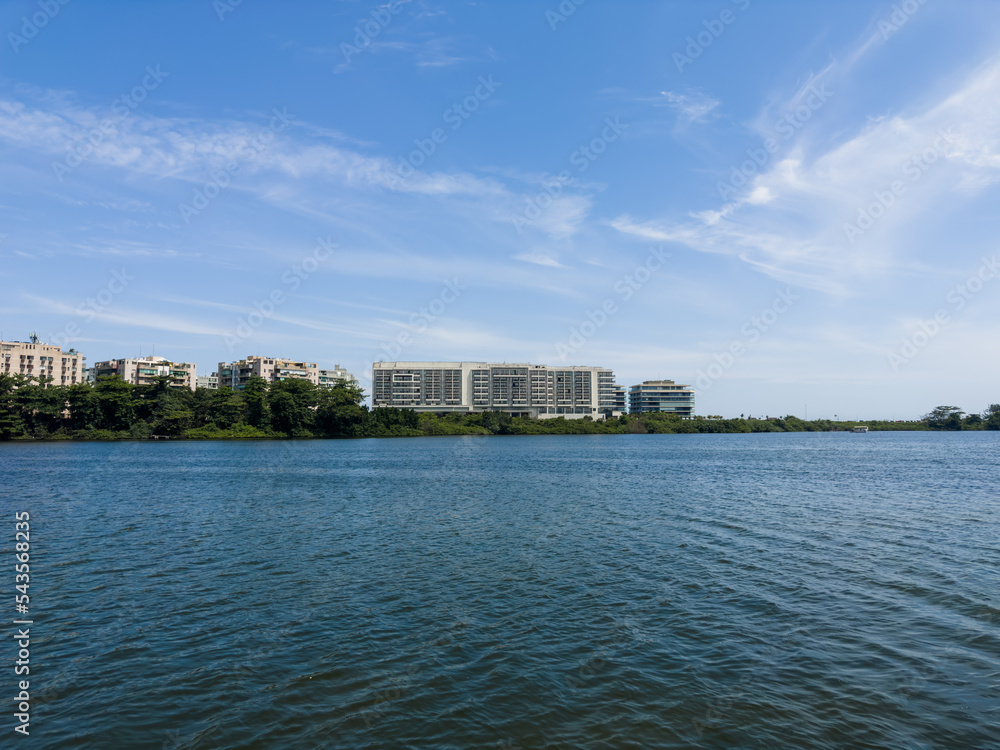 View of the Marapendi lagoon with buildings in the background and surrounding vegetation and trees. Hills in the background. Located near Praia da Reserva in Rio de Janeiro