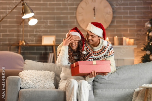 Fotografi Young man surprising his beloved girlfriend with present at home on Christmas ev