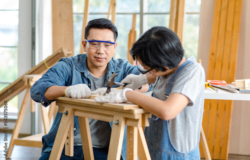 Asian professional carpenter engineer dad teaching young boy son in jeans outfit with gloves safety goggles nailing steel hammer on wood stick on workbench in home housing building construction site
