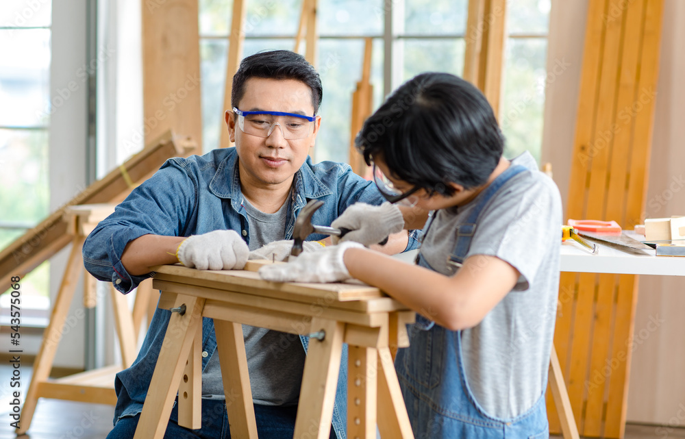 Asian professional carpenter engineer dad teaching young boy son in jeans outfit with gloves safety goggles nailing steel hammer on wood stick on workbench in home housing building construction site Asian professional carpenter engineer dad teaching young boy son in jeans outfit with gloves safety goggles nailing steel hammer on wood stick on workbench in home housing building construction site