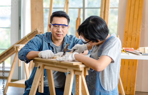 Asian professional carpenter engineer dad teaching young boy son in jeans outfit with gloves safety goggles nailing steel hammer on wood stick on workbench in home housing building construction site