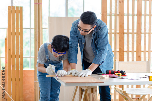 Asian professional male carpenter woodworker engineer dad in jeans outfit with safety gloves goggles helping teaching young boy son using handsaw cutting wooden stick in housing construction site