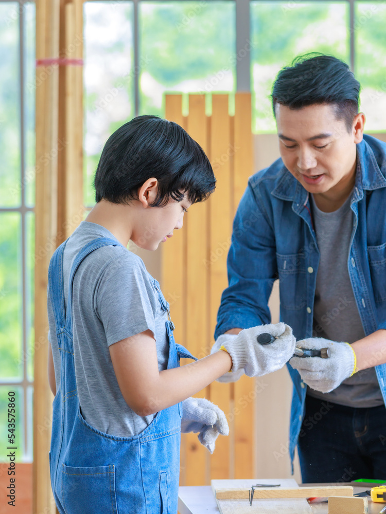 Obraz premium Asian cheerful male dad son carpenter woodworker colleague in jeans outfit with safety gloves helping using pencil drawing writing blueprint on working workshop table in housing construction site