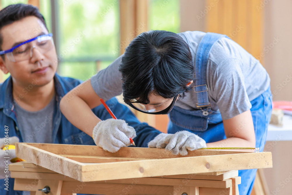 Asian male carpenter woodworker engineer dad in jeans outfit with safety gloves and glasses goggles helping teaching son using pencil marking wood stick board plank on workbench in construction site Asian male carpenter woodworker engineer dad in jeans outfit with safety gloves and glasses goggles helping teaching son using pencil marking wood stick board plank on workbench in construction site