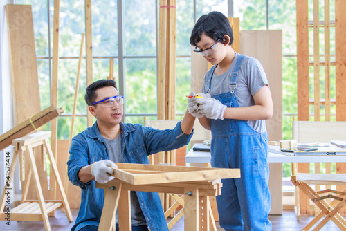 Asian professional carpenter engineer dad teaching young boy son in jeans outfit with gloves safety goggles in home housing building construction site.